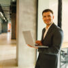 Smiling man in a suit holding a laptop in a modern office hallway.