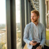 A smiling man in a blazer is sitting by the window of a modern office with a smartphone, looking out at the city.