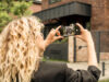 A woman with blonde curls is taking a photo of a modern brick apartment building with her smartphone.
