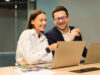 A woman and a man are sitting at a table in an office, smiling as they look at something together on a laptop.
