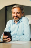 A smiling man in a light blue shirt is sitting at a desk, looking at his smartphone.
