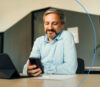A smiling man in a light blue shirt is sitting at a desk, looking at his smartphone.